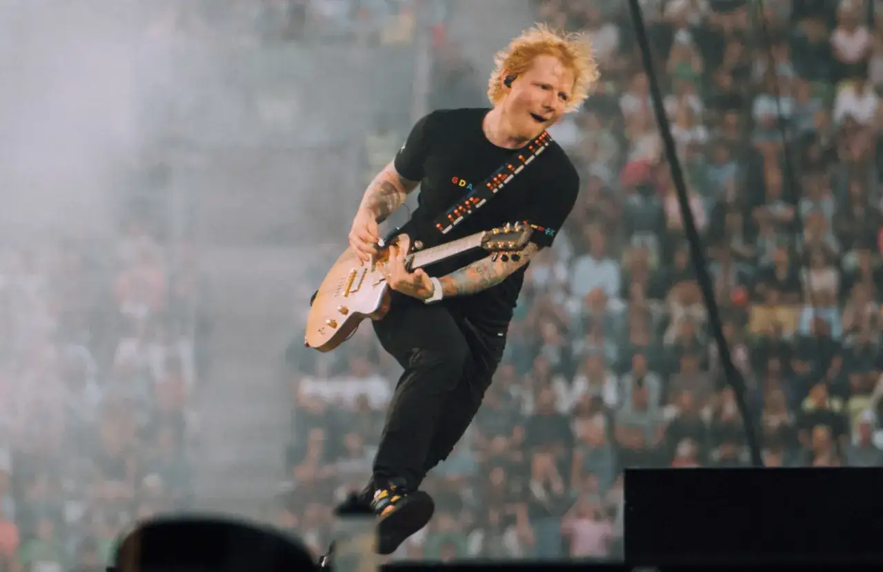 Ed Sheeran skacze na scenie, grając na gitarze, w tle tłum fanów.