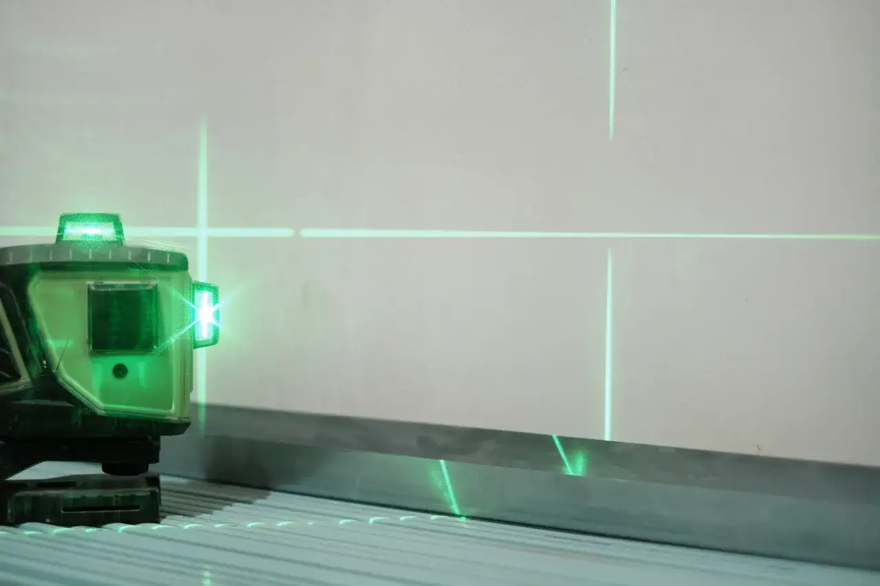 Zielona poziomica laserowa emituje linie na ścianie i metalowym profilu.