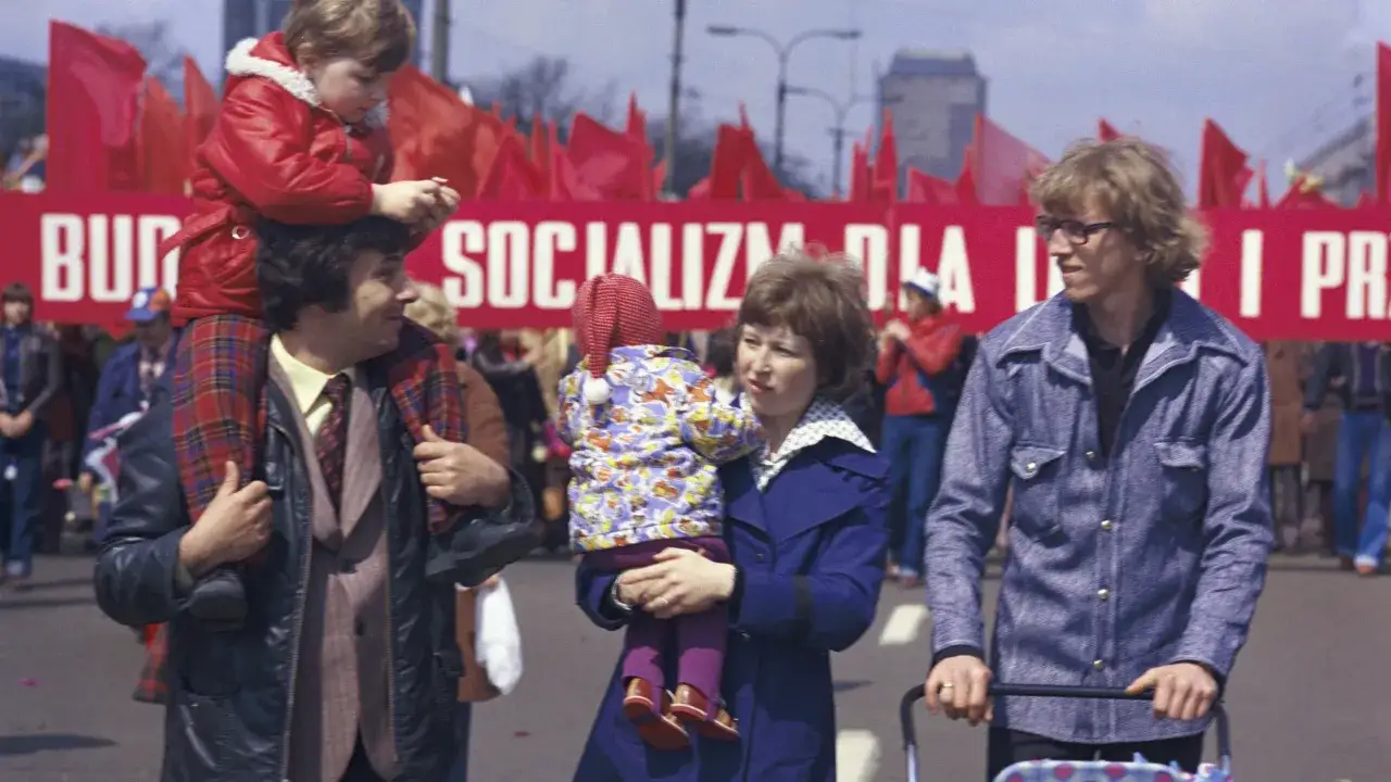 Rodzina na tle czerwonego transparentu z napisem "SOCJALIZM DLA PRACY" i czerwonych flag.