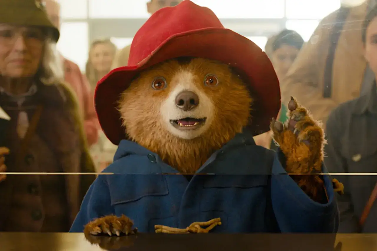 Pluszowy miś Paddington w czerwonym kapeluszu i niebieskim płaszczu, machający łapką.