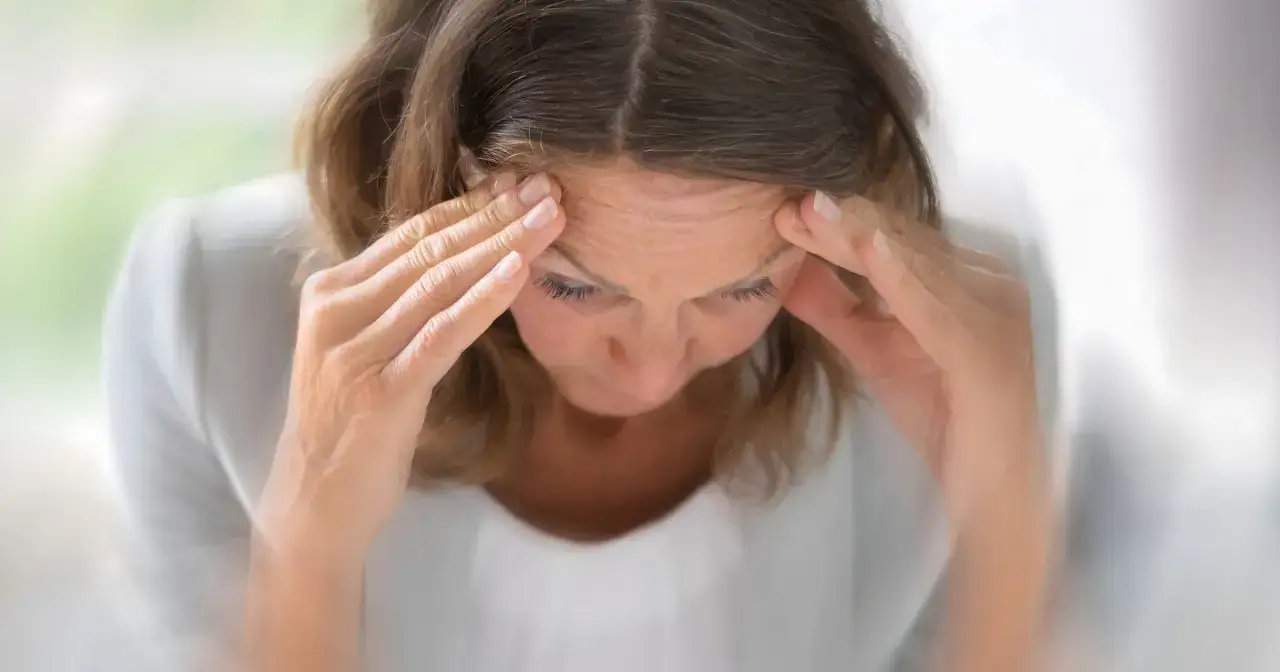 Kobieta z dłońmi na skroniach, wyraźnie cierpiąca z powodu bólu głowy, być może po przejściu udaru.
