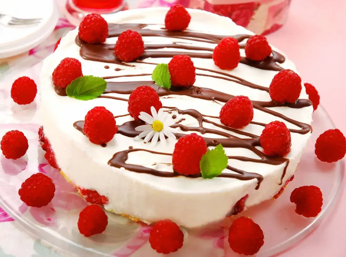 Malinowa rozkosz bez pieczenia: biały tort udekorowany świeżymi malinami, czekoladą i listkami mięty.