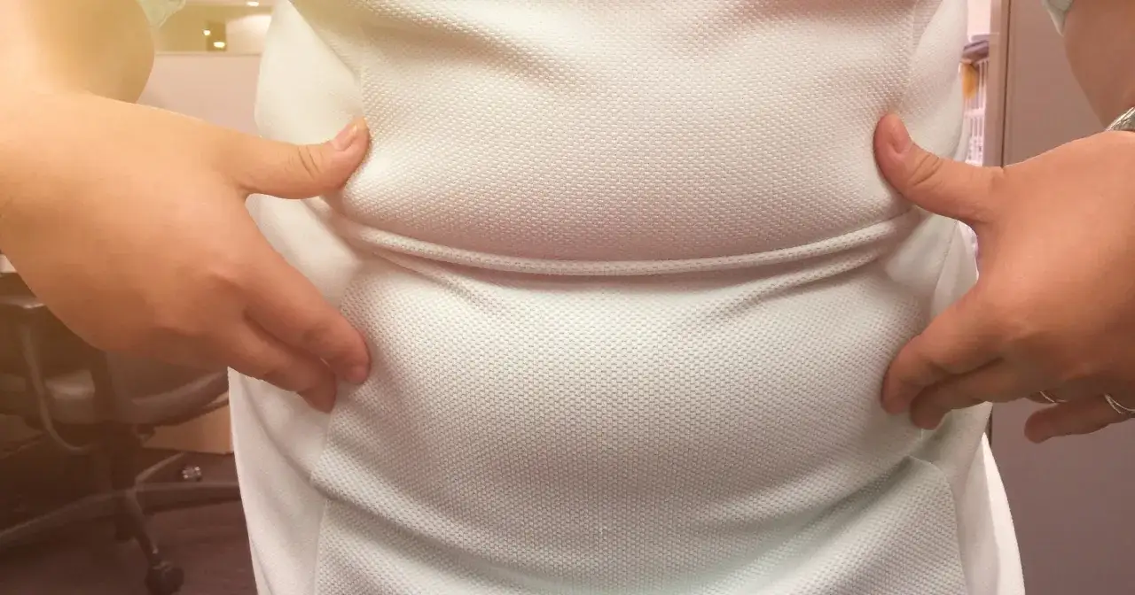 Dłonie uciskają białą koszulkę, uwydatniając wzdęty brzuch. Przyczyny mogą być różne, od diety po stres.