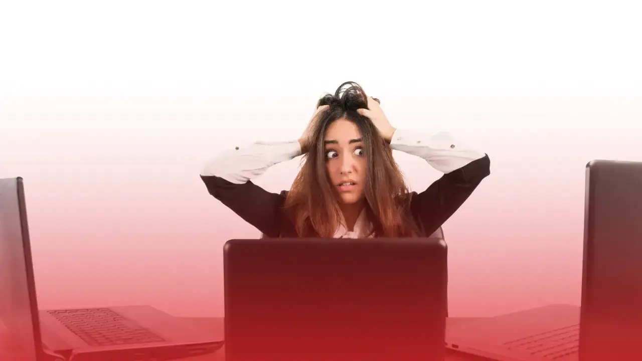 Kobieta z przerażeniem w oczach, rękami wplątanymi we włosy, siedzi przy biurku otoczona laptopami. To obraz stresu w pracy.
