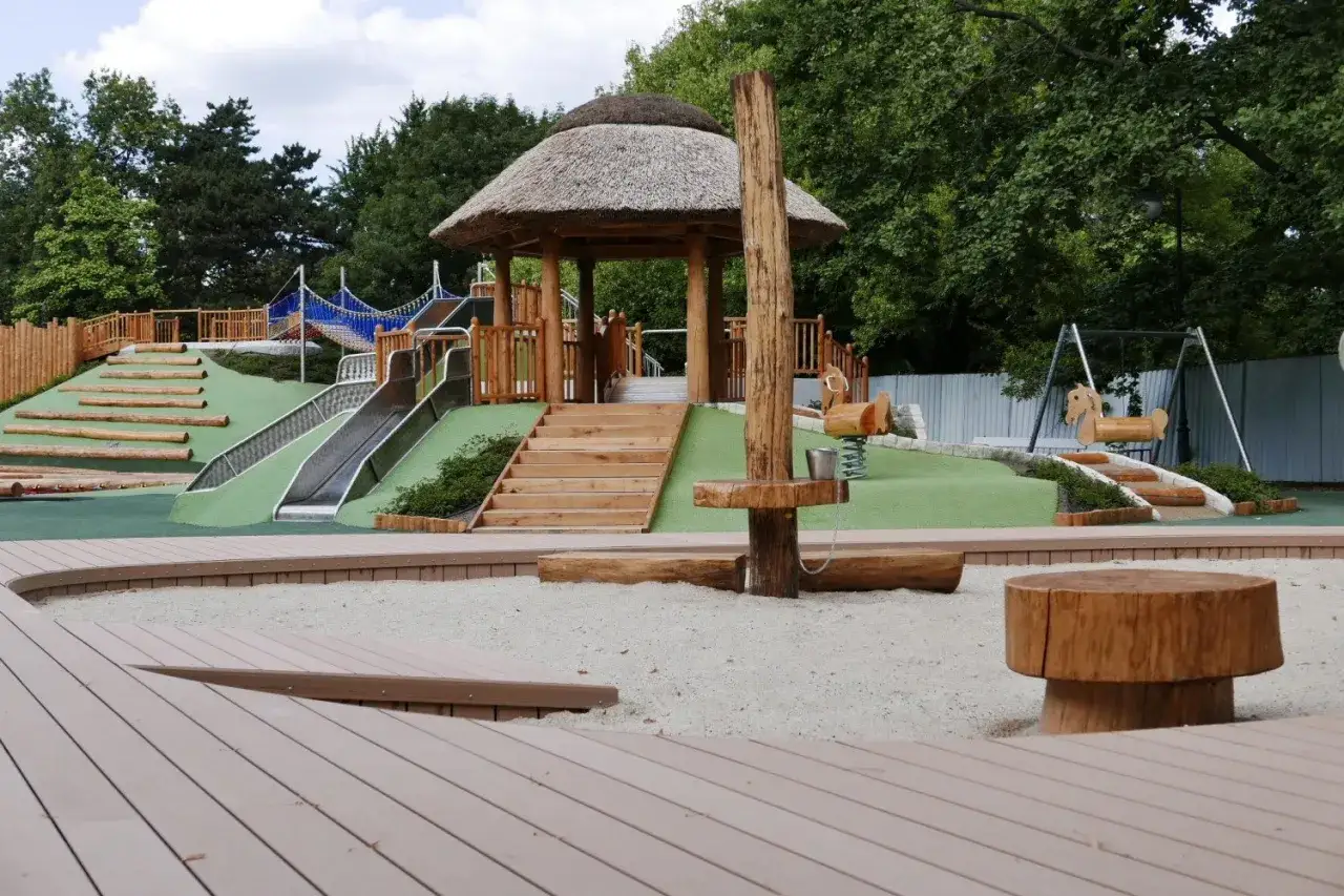 Nowoczesny plac zabaw z drewnianymi elementami, zjeżdżalniami i domkiem pod strzechą.