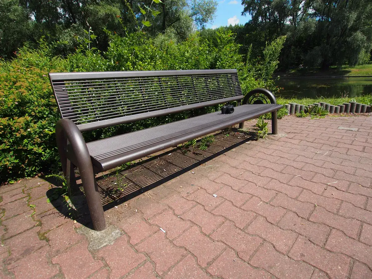 Szeroki kadr ukazuje pustą, metalową ławkę w parku, obok której leży aparat fotograficzny. W tle zielone drzewa i staw.