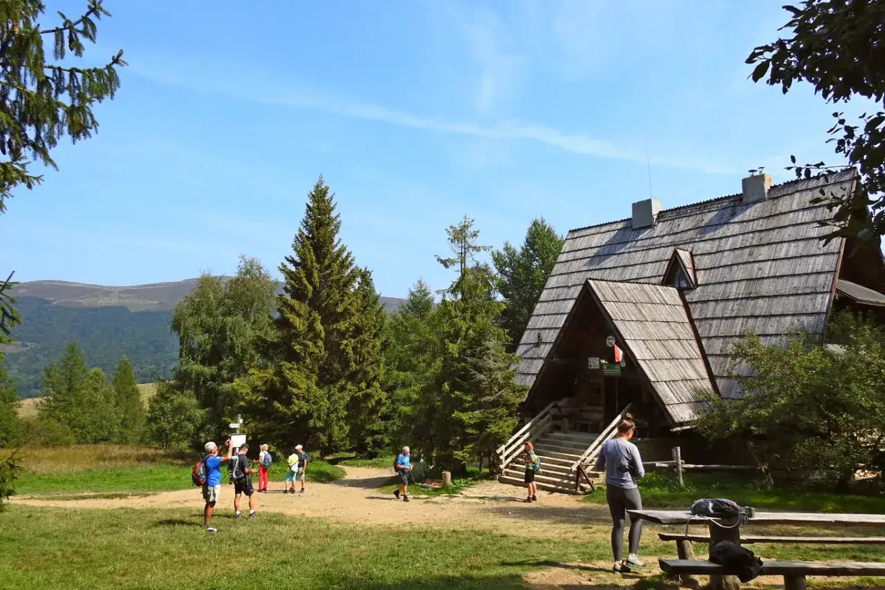 Bacówka PTTK pod Małą Rawką, drewniany schron turystyczny w otoczeniu zieleni, z kilkoma turystami na szlaku.