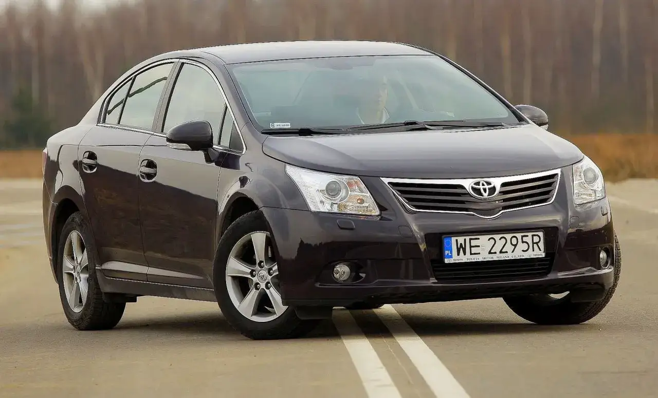 Ciemny sedan Toyota Avensis na drodze. Kierowca za kierownicą. Szczegółowe dane techniczne tego modelu są dostępne online.