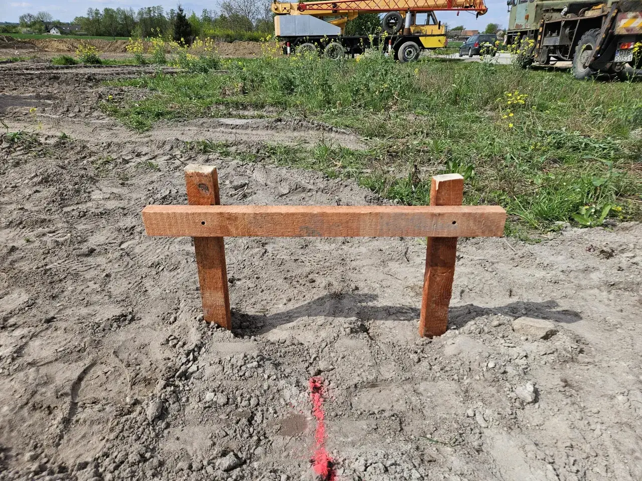 Drewniany palik z poprzeczką wbity w ziemię na budowie, z zaznaczoną czerwoną linią.