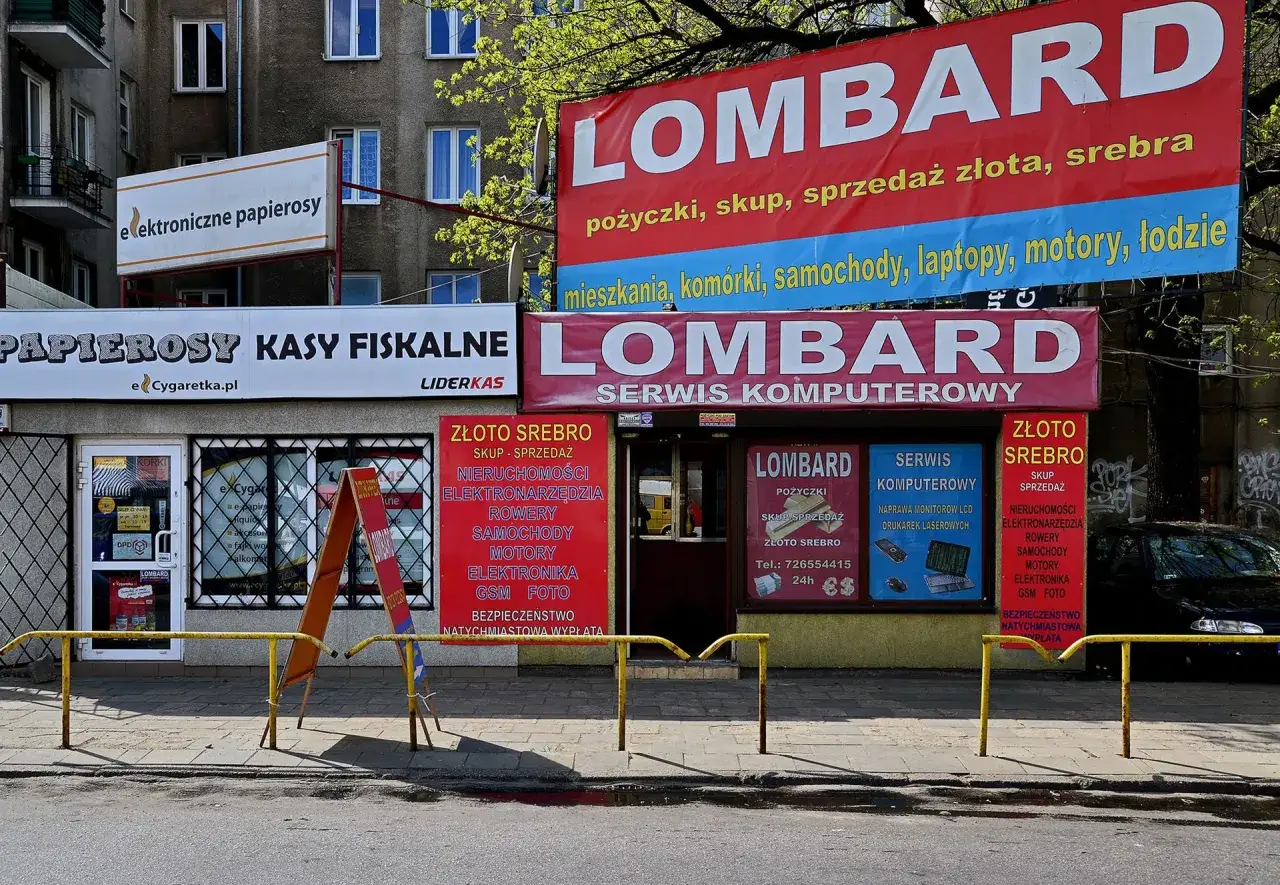 Szyldy lombardu i sklepów z elektroniką i papierosami na tle budynku mieszkalnego.