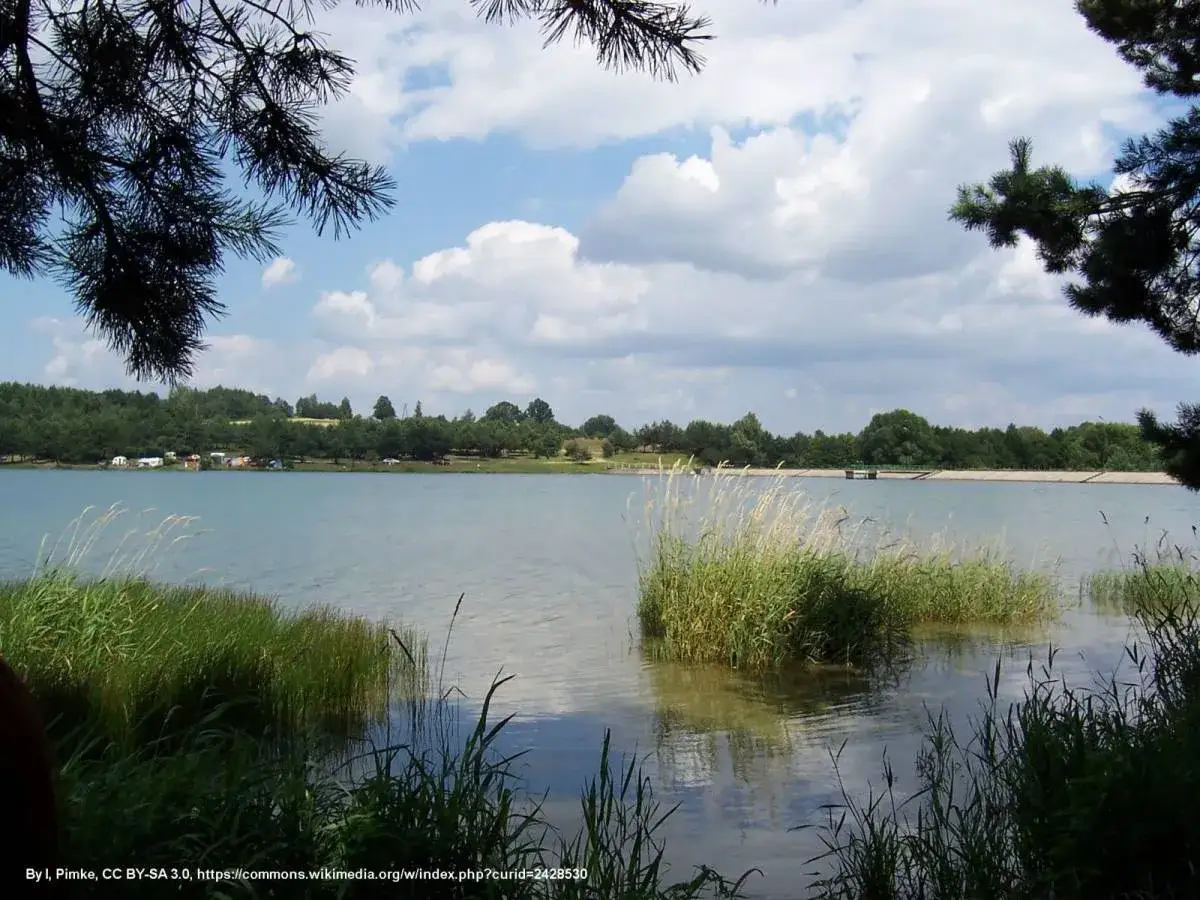 Spokojny zalew Dźibice otoczony zielenią, z kempingiem na brzegu i chmurami na niebie.