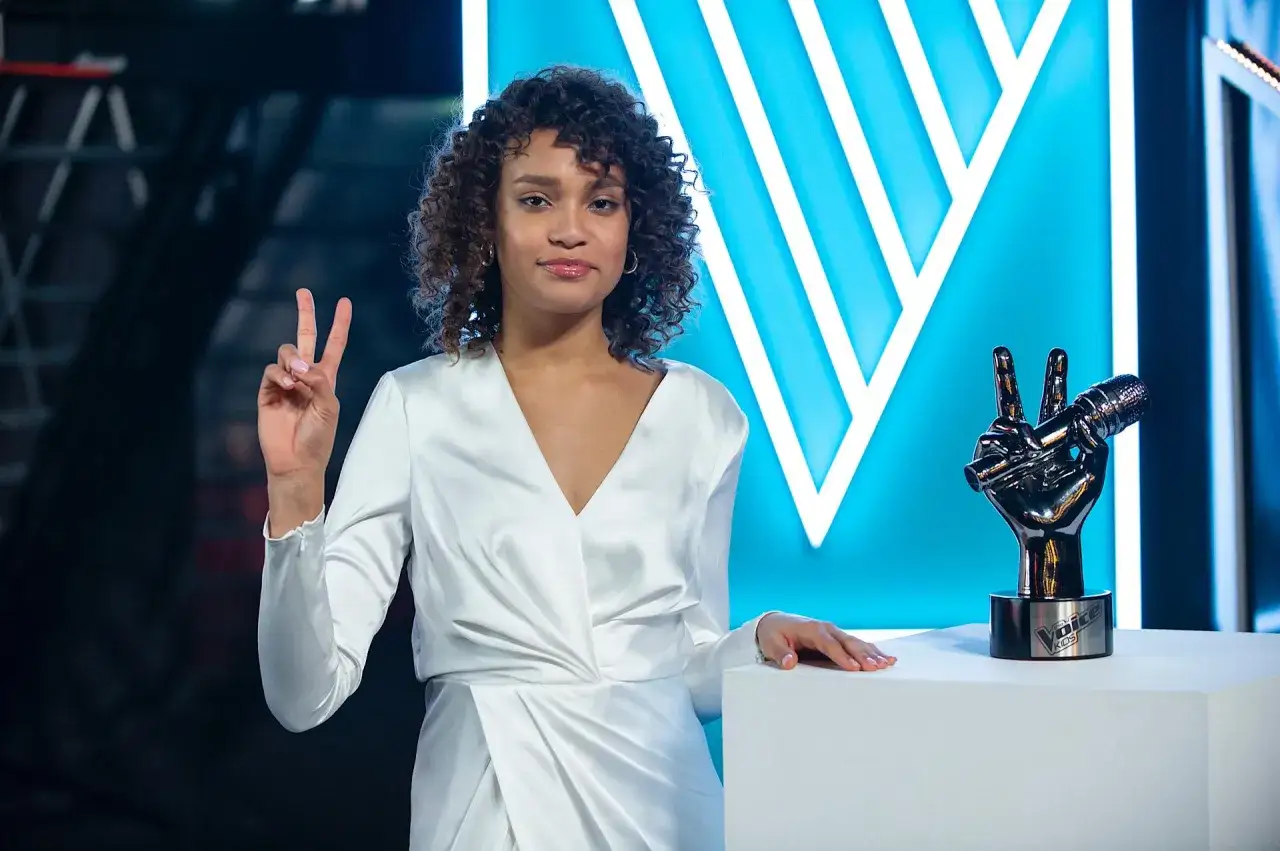 Młoda artystka w białej sukni z trofeum "The Voice Kids" w dłoni, symbolizującym zwycięstwo.