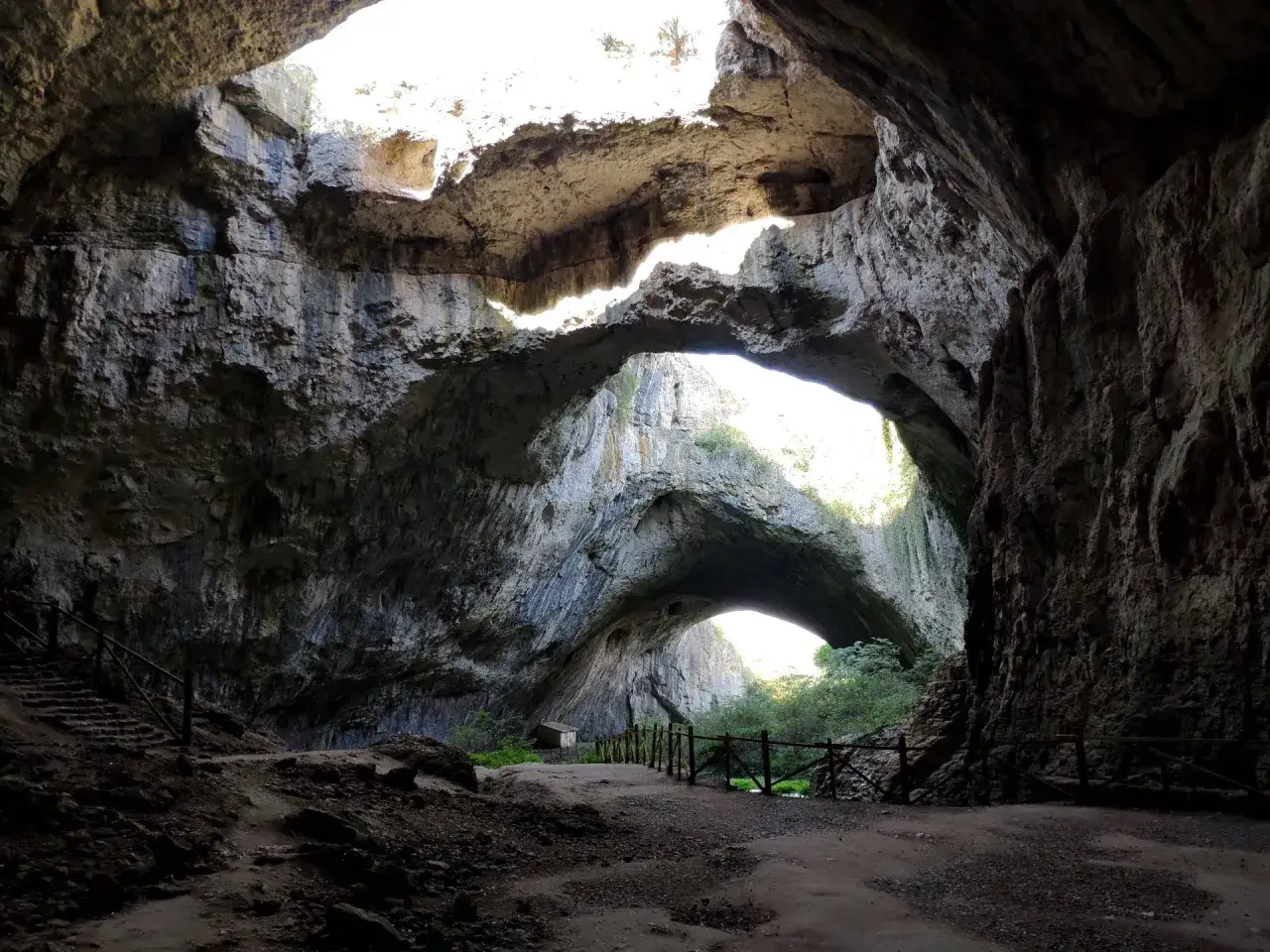 Wnętrze jaskini Devetashka z łukami skalnymi i światłem wpadającym z góry. Ścieżka i schody prowadzą przez ciemne, skaliste wnętrze.