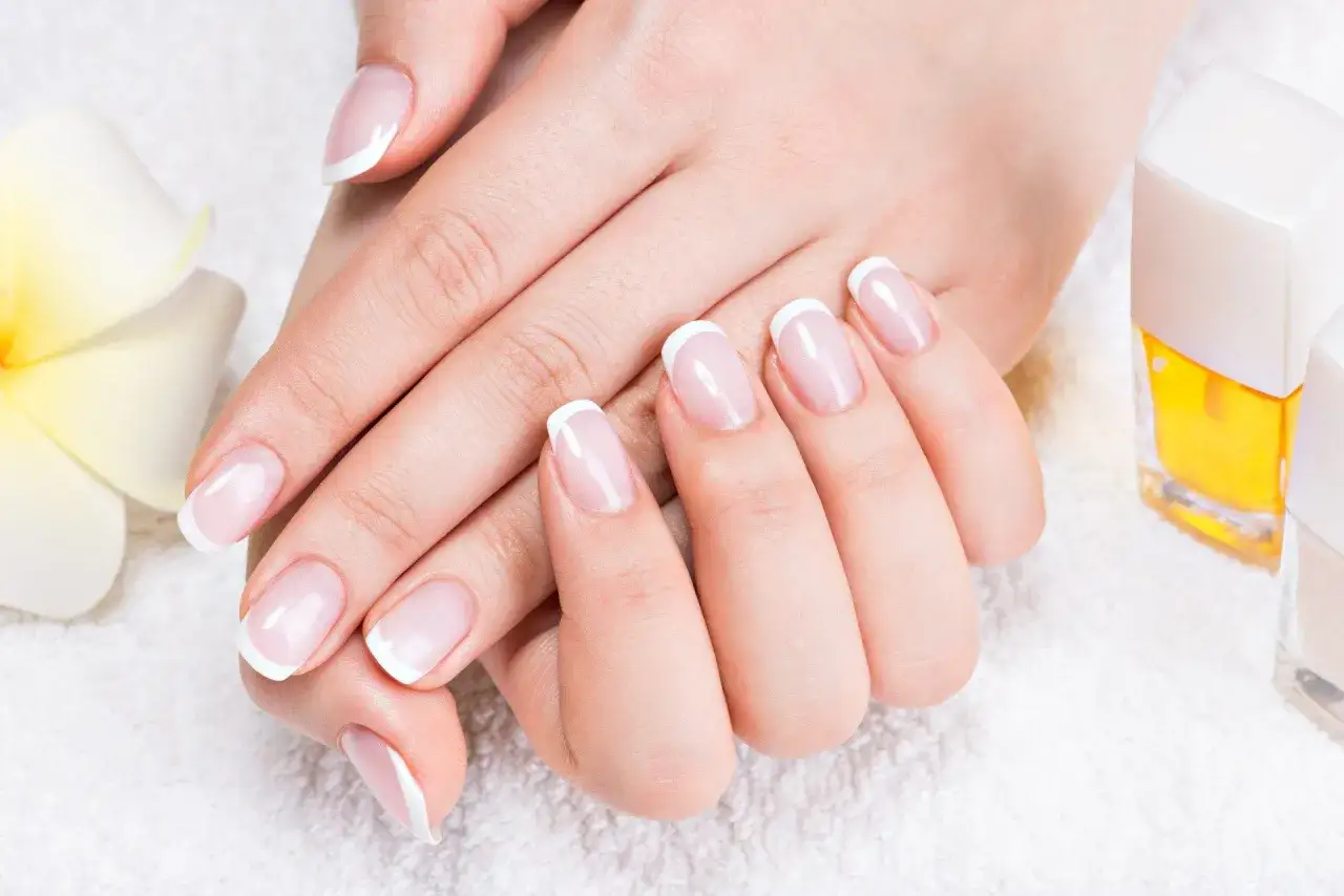 Elegancki manicure french z diamencikami, dłonie zadbane i wypielęgnowane, obok kwiat i lakiery.