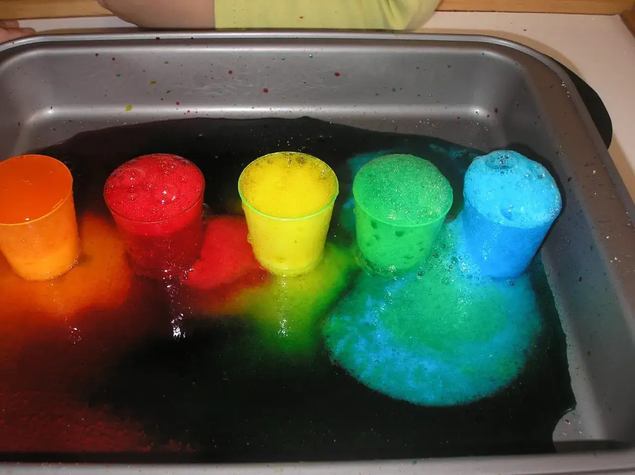 Kolorowe kubeczki z pianą w tacy, tworzące tęczę z płynów.