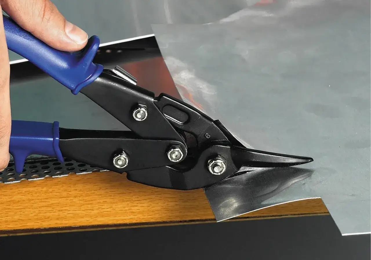 Dłoń trzyma niebieską rękojeść nożyc do blachy, które tną arkusz metalu.