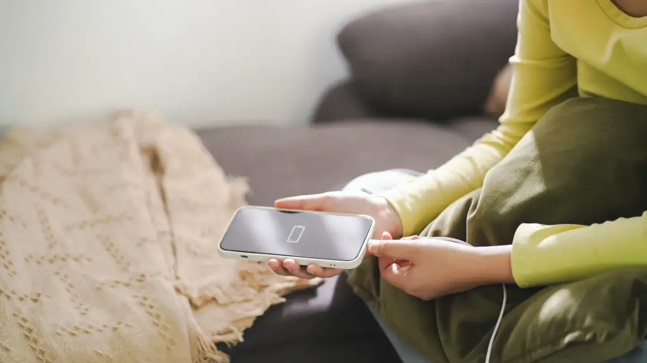 Osoba podłącza iPhone'a do ładowania, co jest kluczowe, jak dbać o baterie iPhone. Niska bateria na ekranie telefonu.