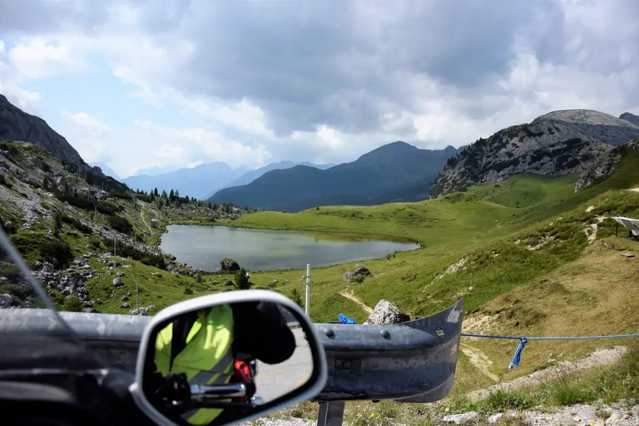 Widok z motocykla na malownicze **przełęcze alpejskie**, jezioro i góry pod zachmurzonym niebem.