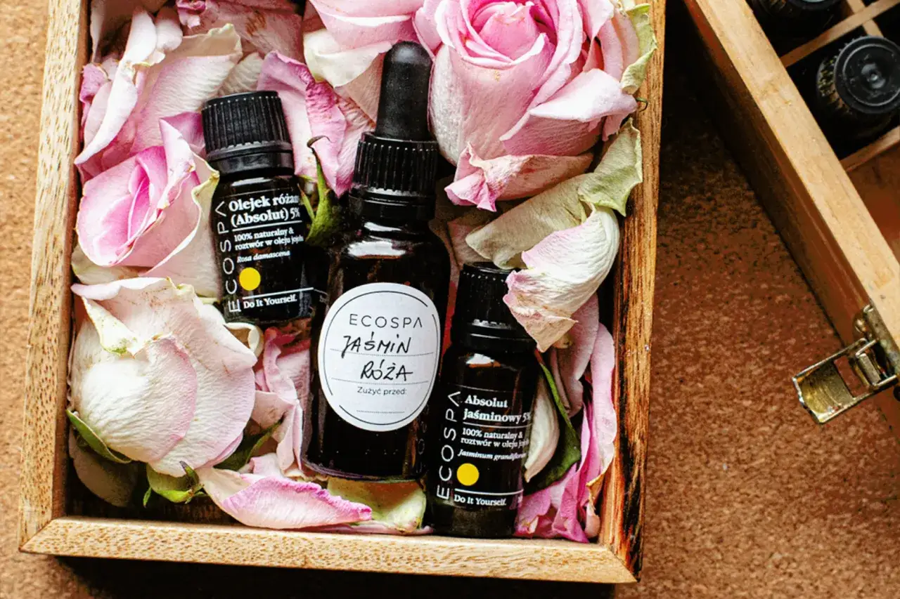 Naturalne olejki różany i jaśminowy w drewnianej skrzynce wśród płatków róż. Dowiedz się, jak oszczędzić na perfumach, wybierając esencje.