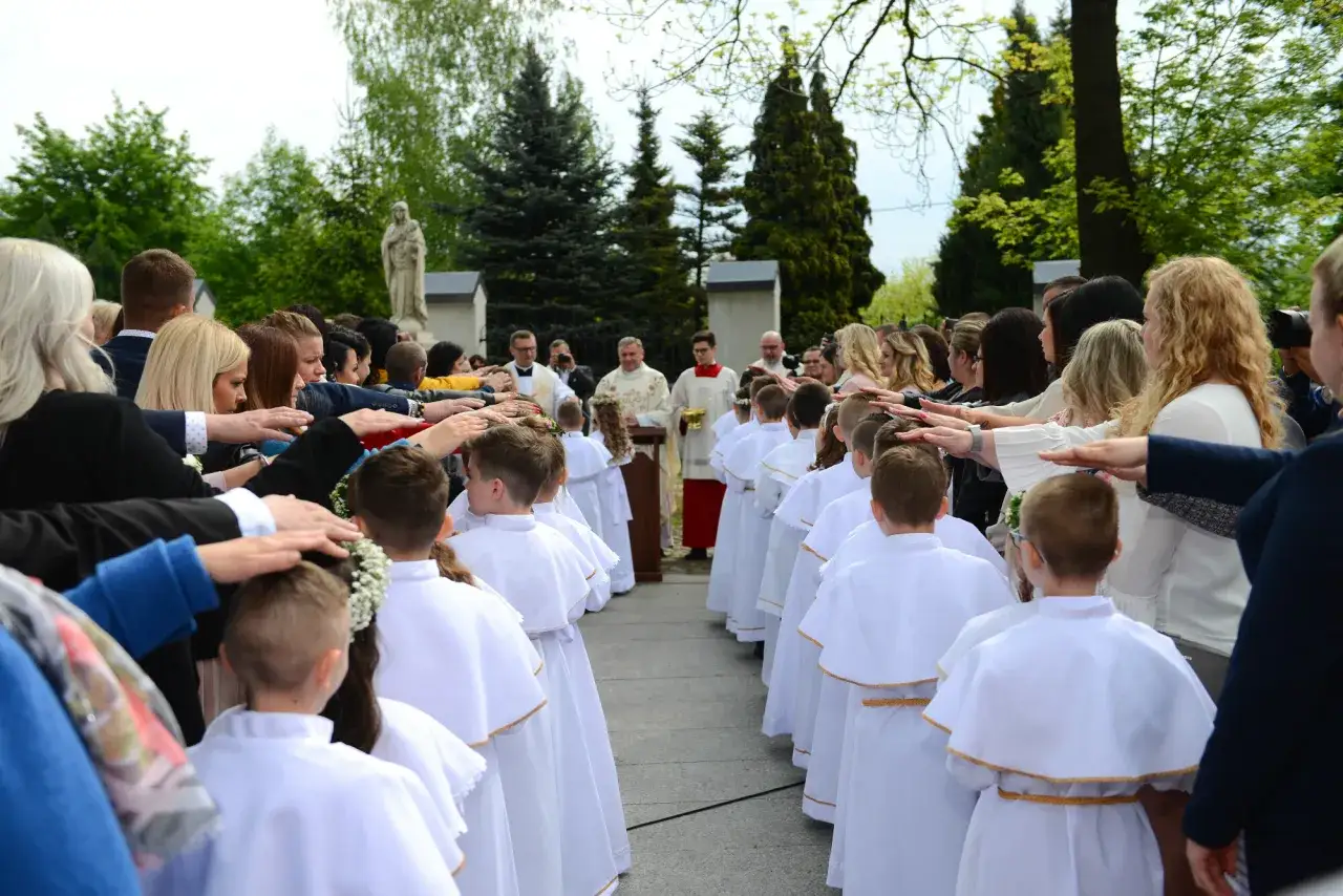 Dzieci w białych strojach komunijnych stoją w rzędzie, a dorośli kładą im ręce na głowach.