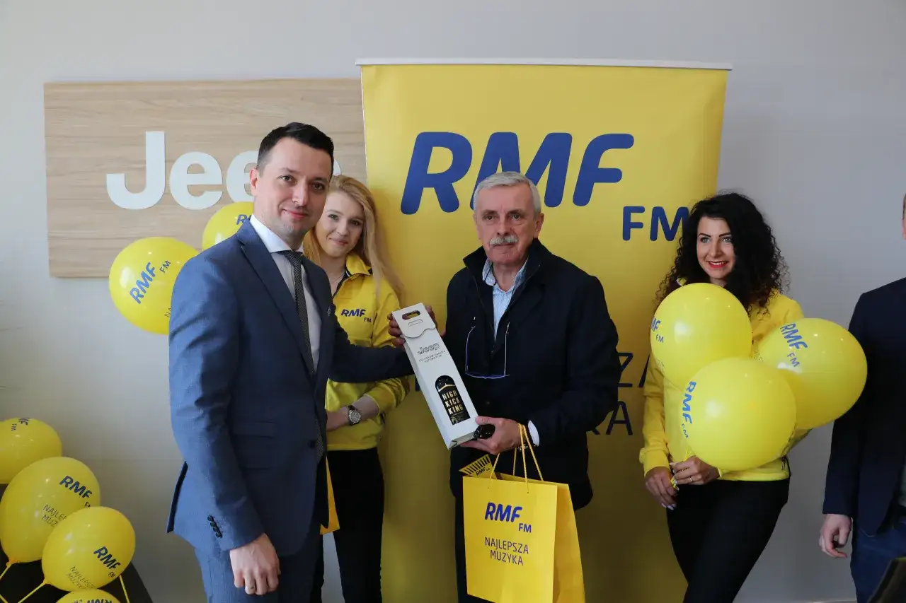 Uroczyste wręczenie nagrody w ramach finału loterii RMF FM. Mężczyzna w garniturze odbiera prezent od uśmiechniętej kobiety w żółtej kurtce.