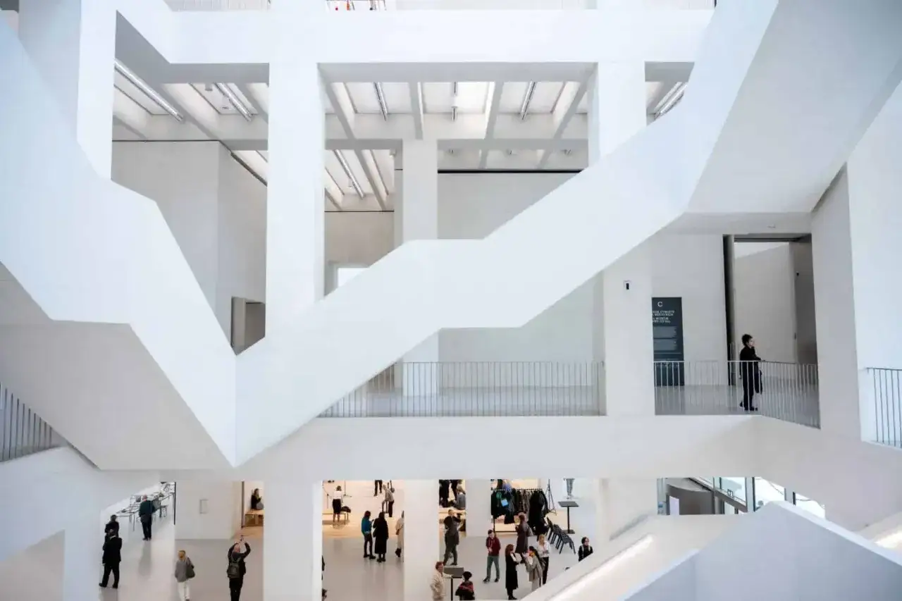 Wnętrze muzeum sztuki nowoczesnej z białymi, geometrycznymi schodami i balustradami. Ludzie zwiedzają, kupując bilety i podziwiając ekspozycje.