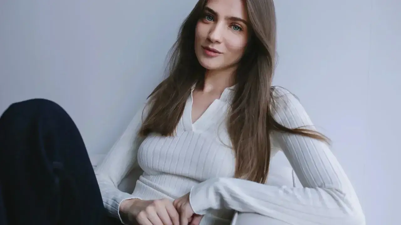 Kobieta o delikatnych rysach i niebieskich oczach, idealny **typ urody lato**. Długie, brązowe włosy opadają na biały, prążkowany sweter.