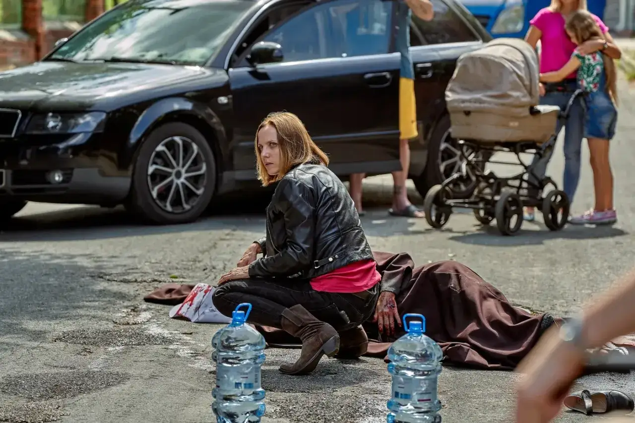 Kobieta w czarnej ramonesce klęczy obok zakrwawionej osoby na drodze. W tle czarny samochód i wózek dziecięcy. Czy to lipowo zmowa milczenia?