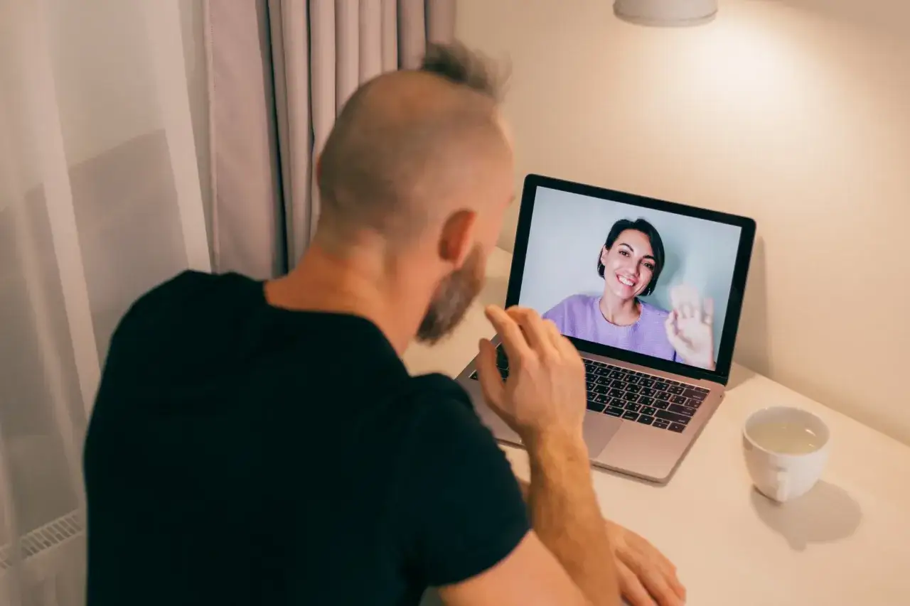 Mężczyzna odbywa konsultację psychologiczną online. Na ekranie laptopa widoczna uśmiechnięta kobieta machająca ręką.
