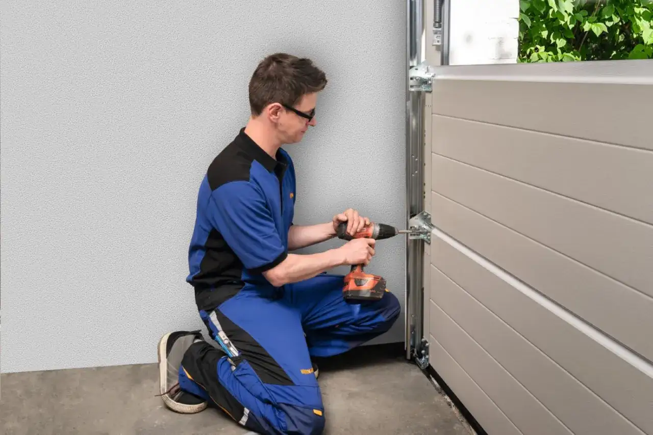 Monter w niebieskim kombinezonie przykręca wkrętarką zawias bramy garażowej.