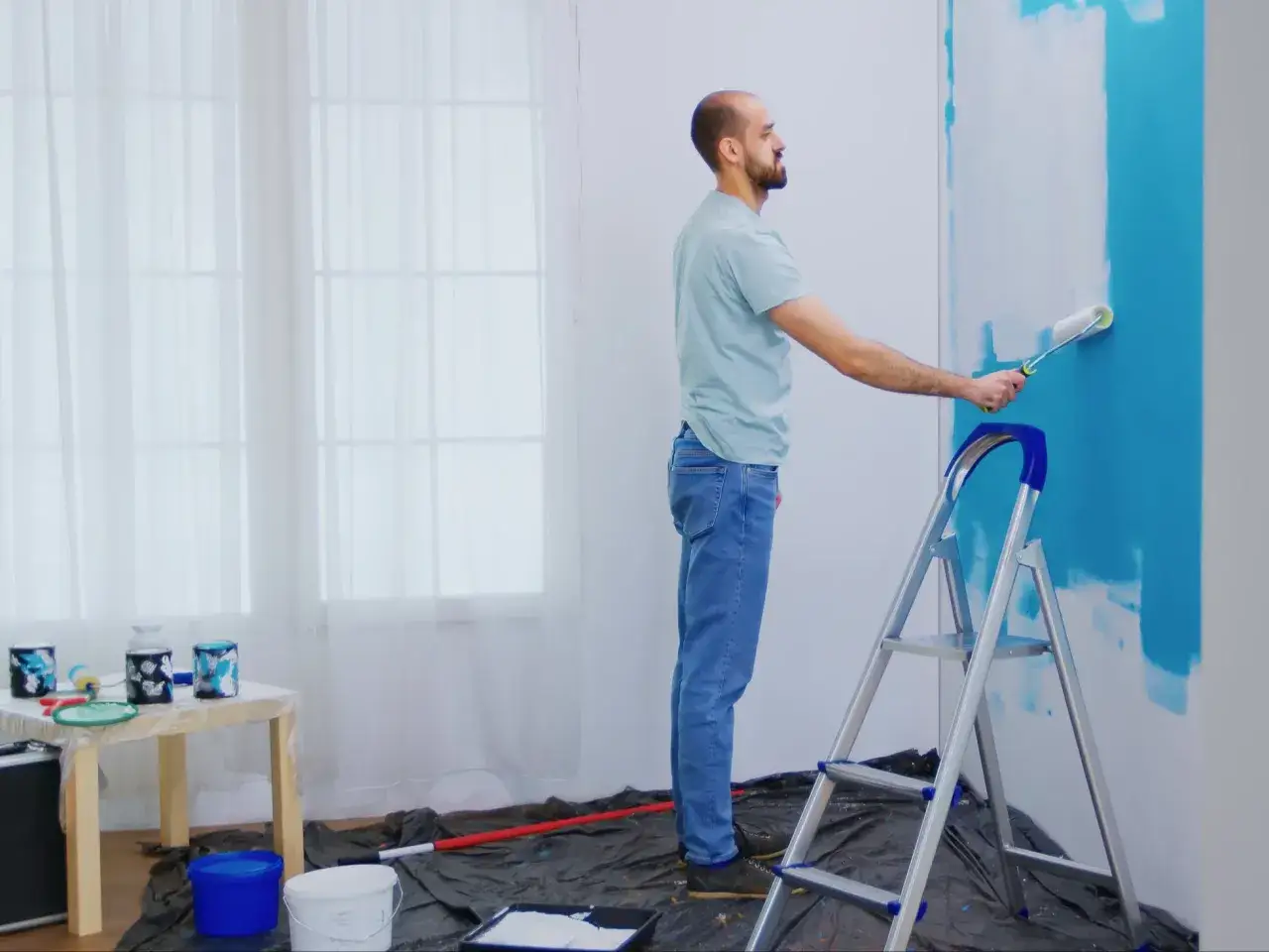 Mężczyzna maluje ścianę na niebiesko wałkiem, stojąc na drabinie.