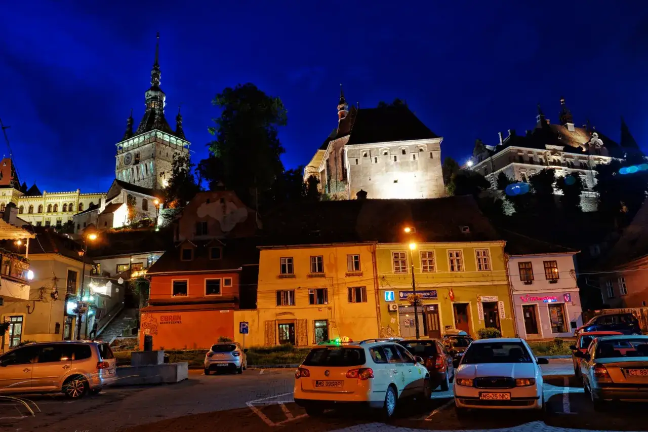 Nocne, oświetlone budynki i wieża zegarowa w Sighișoara. Atrakcje miasta nocą, z samochodami na pierwszym planie.