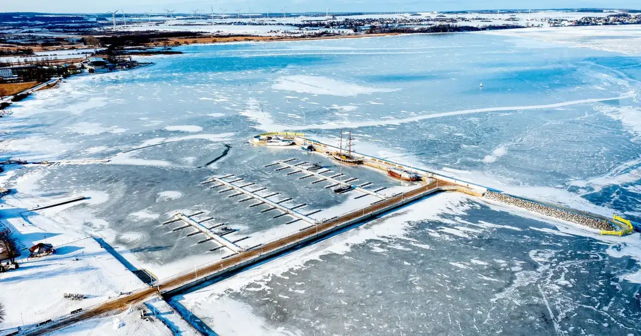 Zamarznięte morze bałtyckie zimą. Widok na zamarzniętą zatokę z przystanią, statkami i wiatrakami na horyzoncie.