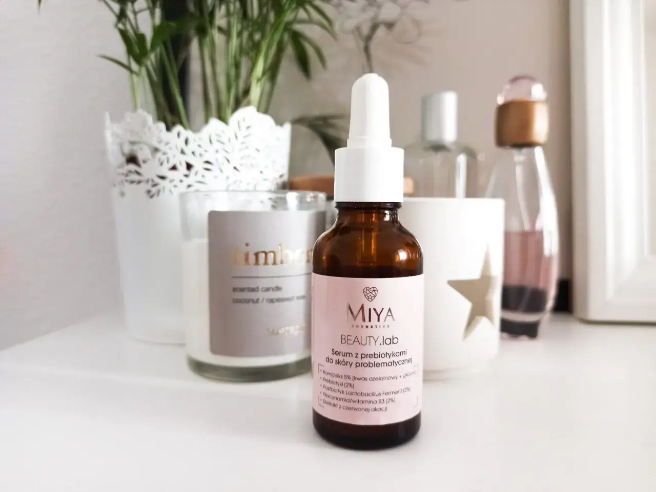 Brązowa buteleczka serum Miya Beauty.lab z pipetką na jasnoróżowej etykiecie, na tle świecy i roślin.