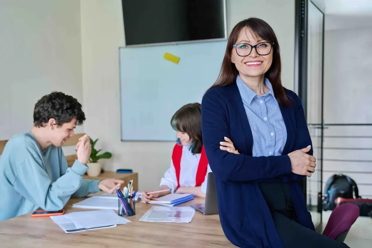 Nauczycielka z uśmiechem nadzoruje zajęcia z wychowawcą. Uczniowie pracują przy biurku, a na tablicy widać rozkład materiału.