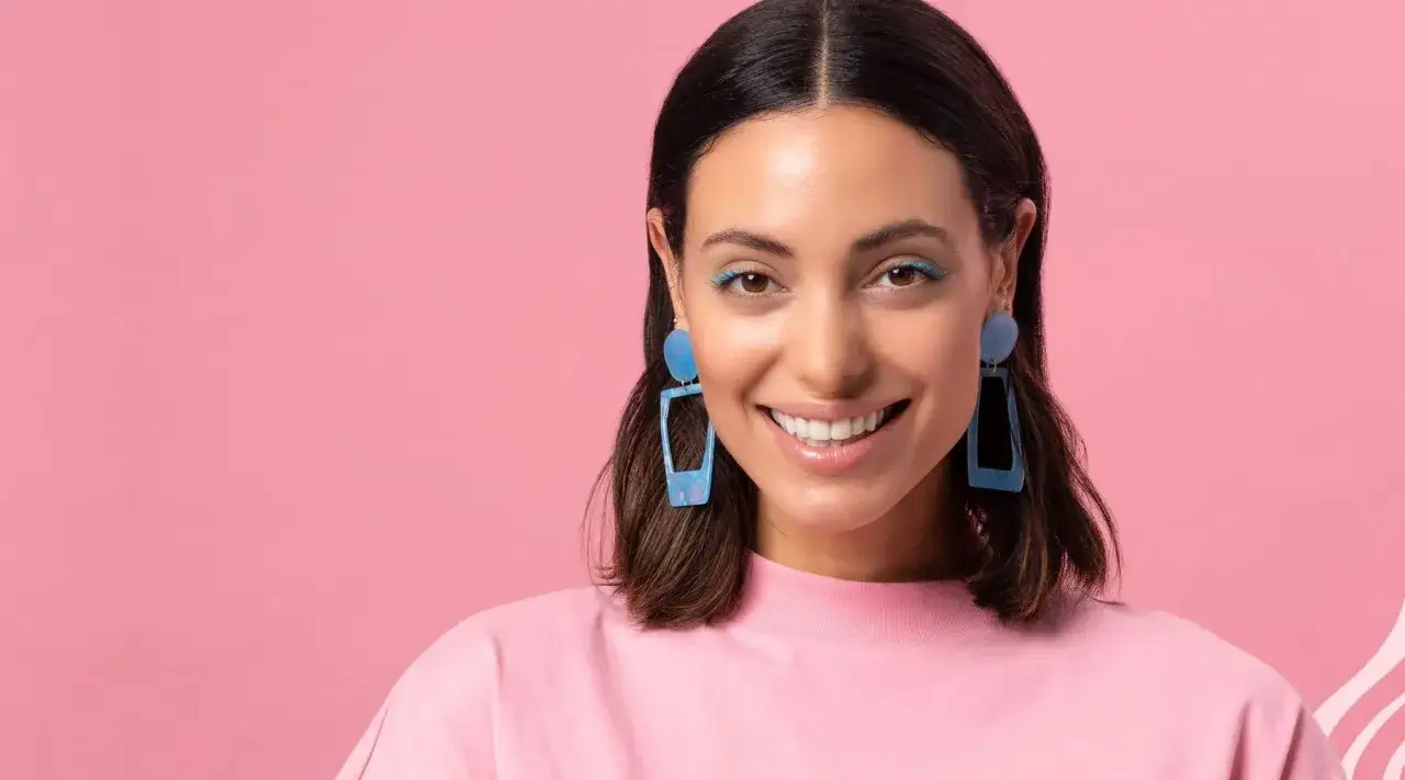 Uśmiechnięta kobieta z niebieskimi kolczykami i makijażem na różowym tle.