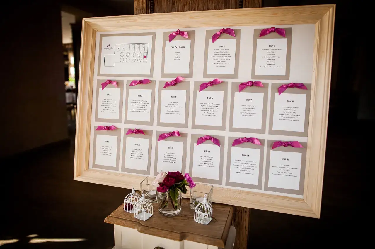 Drewniana rama z planem stołów weselnych i listą gości, ozdobiona różowymi wstążkami.