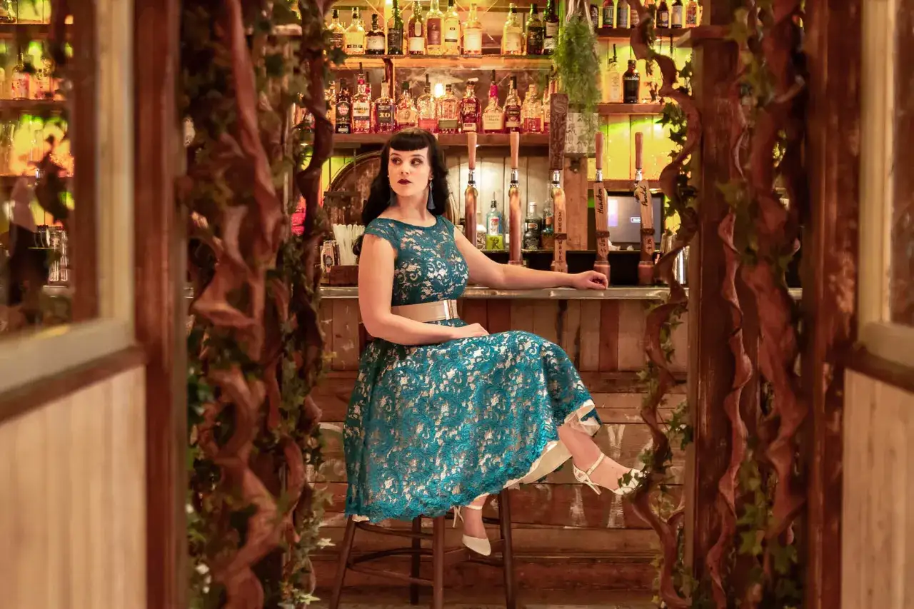Kobieta w turkusowej sukience z koronki siedzi przy barze, opierając się o blat.