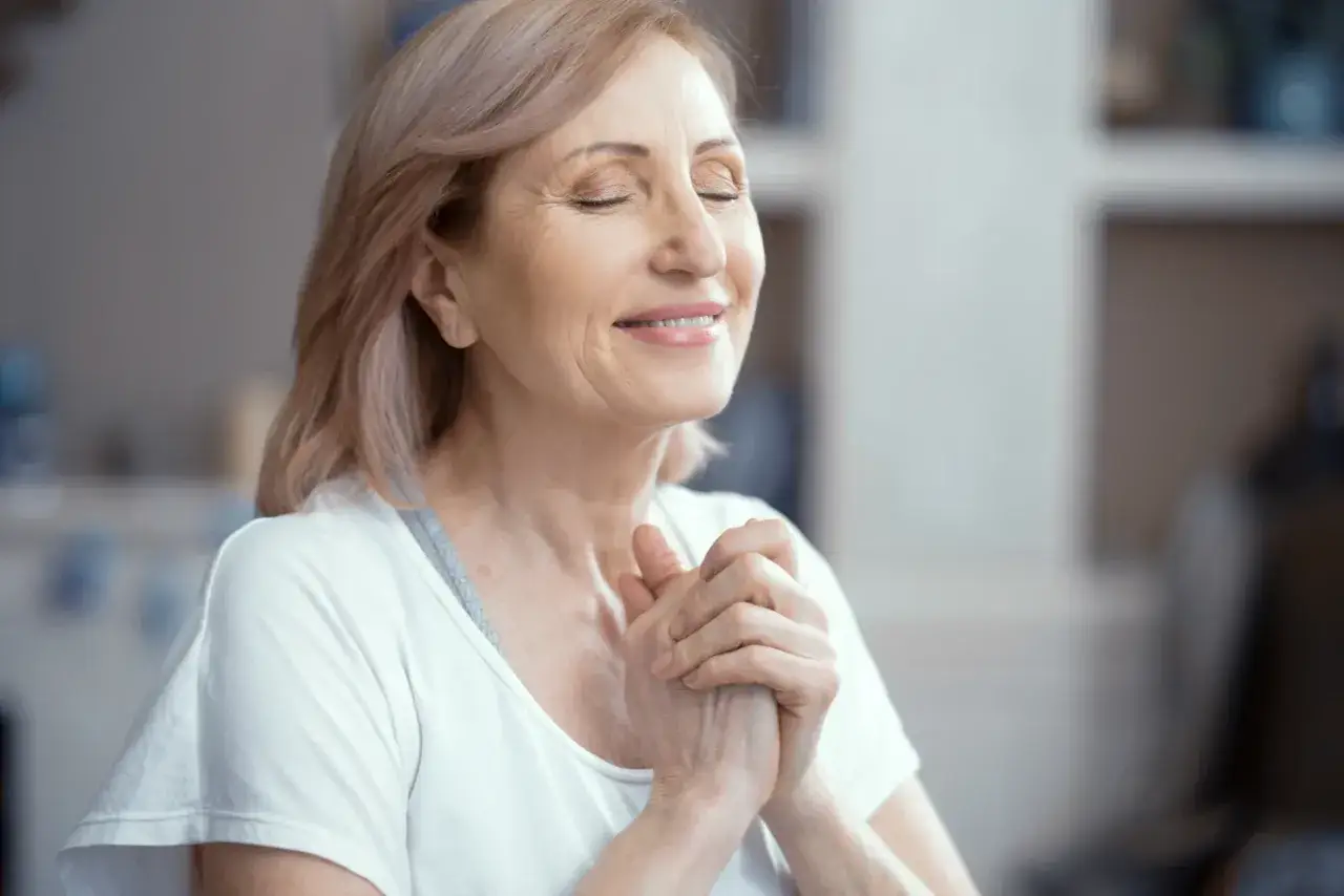 Uśmiechnięta kobieta z zamkniętymi oczami, dłonie złożone na piersi, praktykuje techniki mindfulness.
