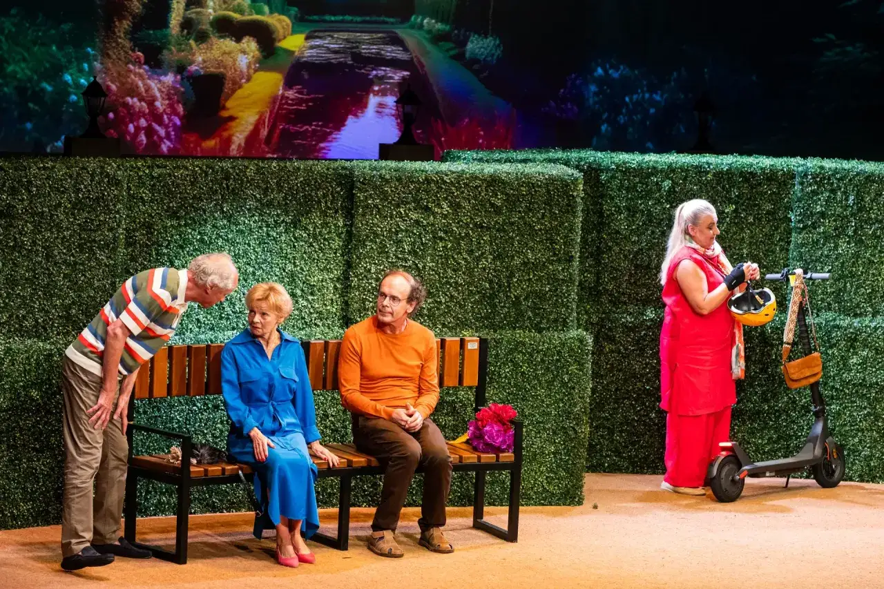 Scena z Teatru Kwadrat w Warszawie. Na ławce siedzą dwie osoby, obok stoi mężczyzna. W tle zielone żywopłoty i ekran z kolorowym obrazem.