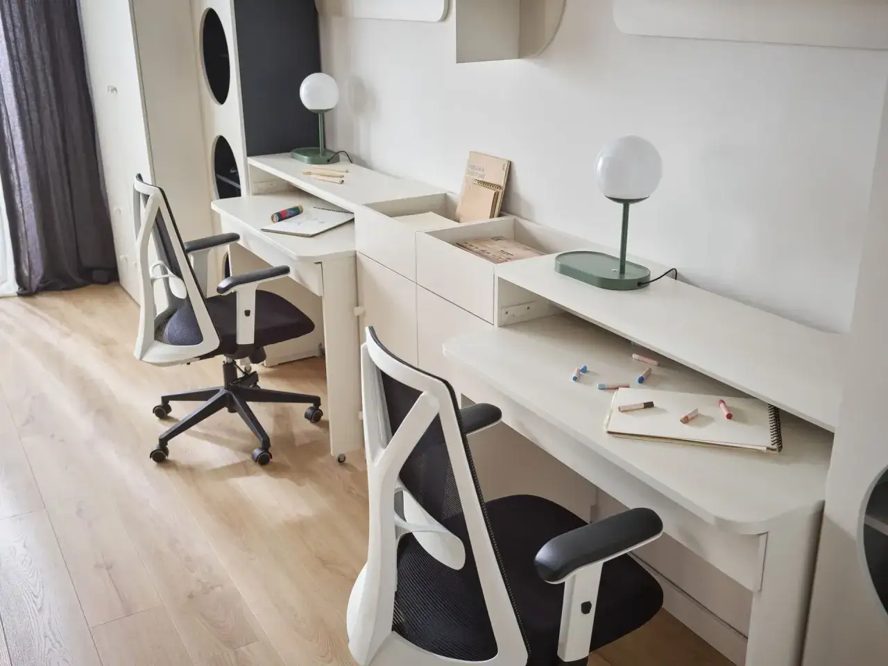 Mały pokój dla 3 dzieci z dwoma biurkami i krzesłami. Na biurkach lampki, zeszyty i kredki.