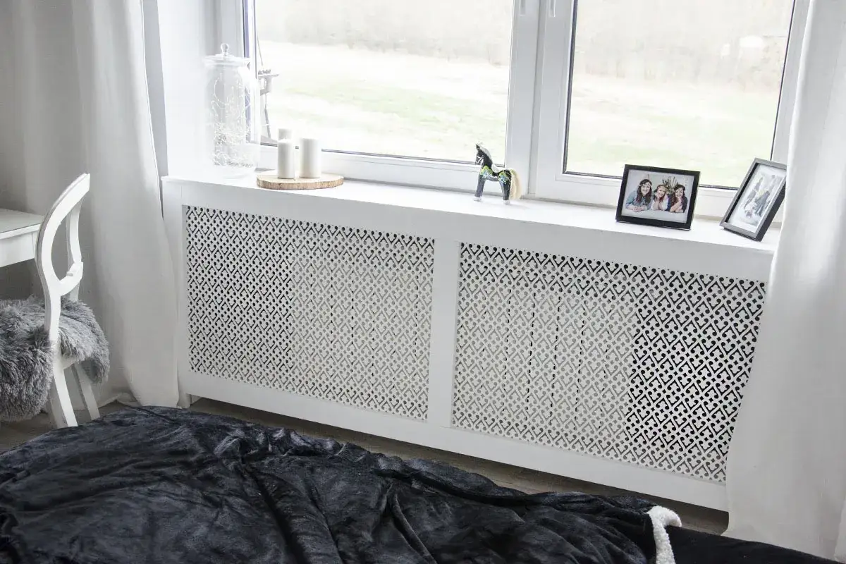 Elegancka osłona kaloryfera z ażurowym wzorem, która stanowi estetyczne rozwiązanie, jak zabudować kaloryfer. Na parapecie zdjęcia i ozdoby.