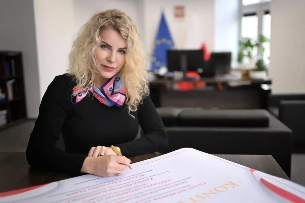 Kobieta z kręconymi blond włosami, w czarnej bluzce i kolorowym szalu, podpisuje dokument. Jest rzecznikiem małych i średnich przedsiębiorców.
