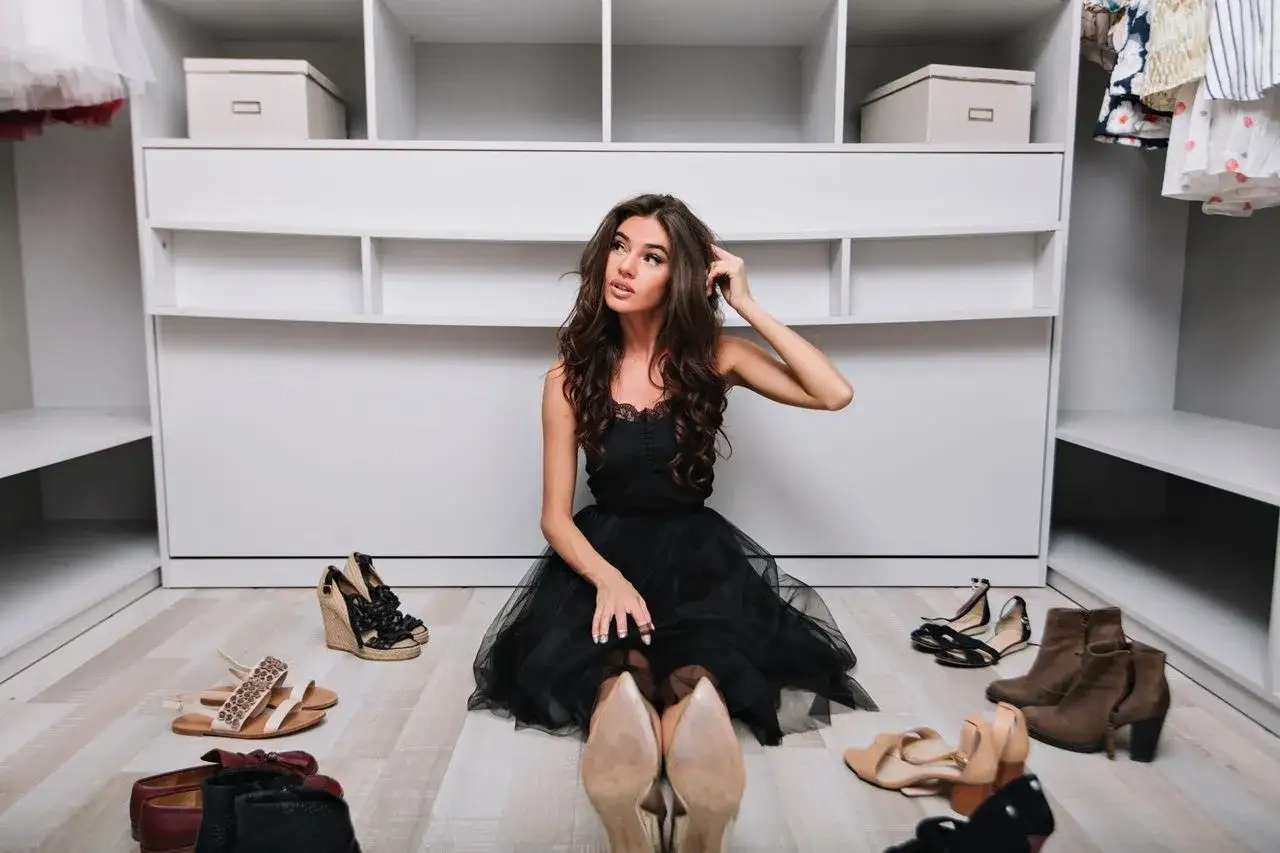 Kobieta w czarnej sukience zastanawia się, jakie buty do szampanskiej sukienki wybrać. Wokół niej rozłożone są różne modele obuwia.