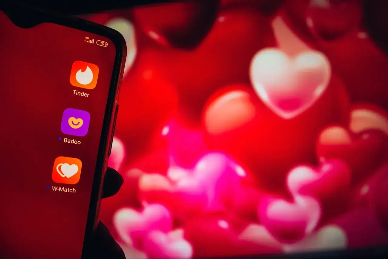 Na ekranie telefonu widać ikony aplikacji randkowych: Tinder, Badoo i W-Match. Tło to czerwone serca, symbolizujące miłość i poszukiwanie tej jedynej osoby. Wybór bezpiecznych portali randkowych to pierwszy krok do znalezienia szczęścia.