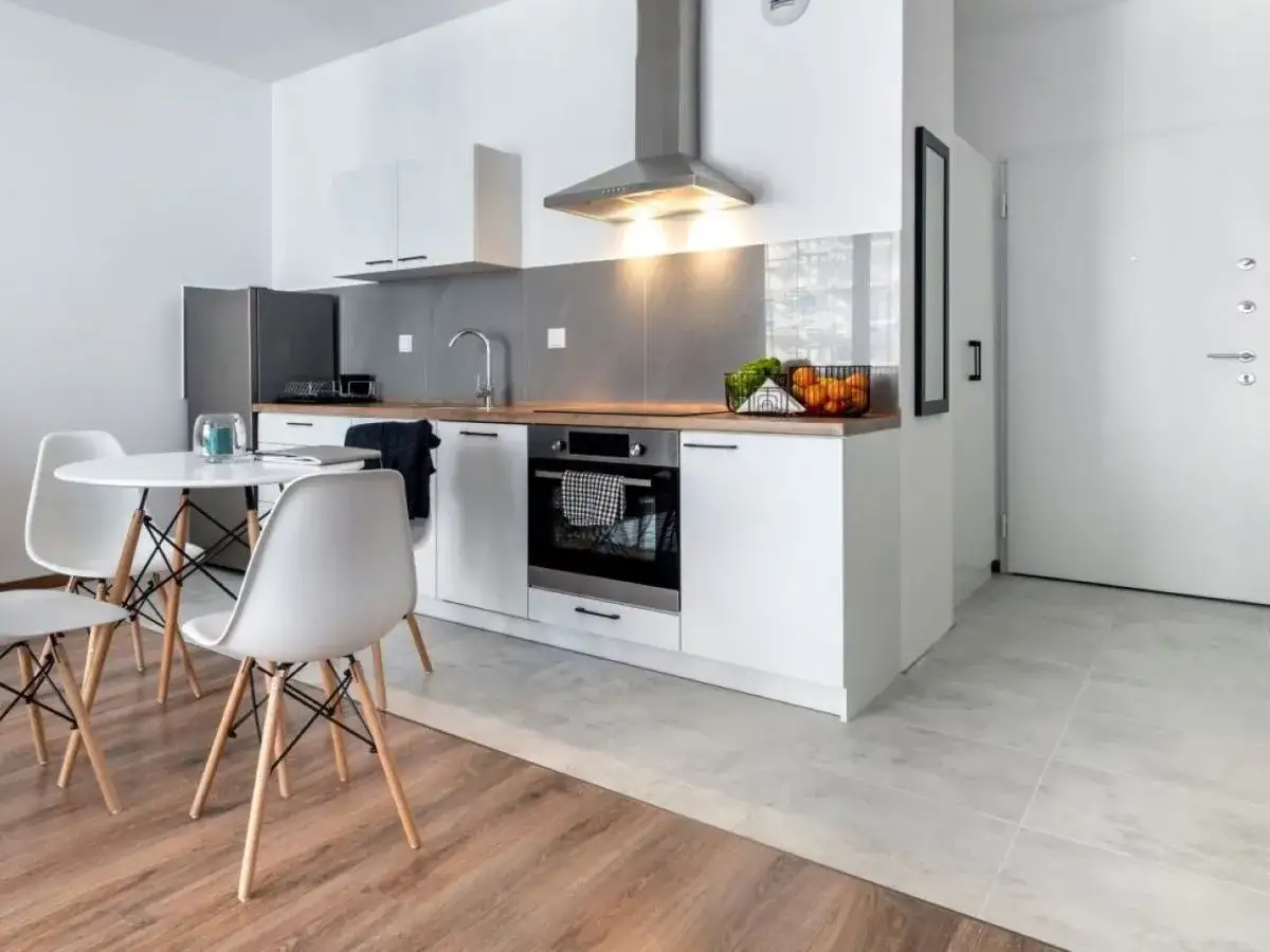 Nowoczesna kuchnia i jadalnia w małym mieszkaniu 35 m. Białe meble, okrągły stół z krzesłami, lodówka i piekarnik.