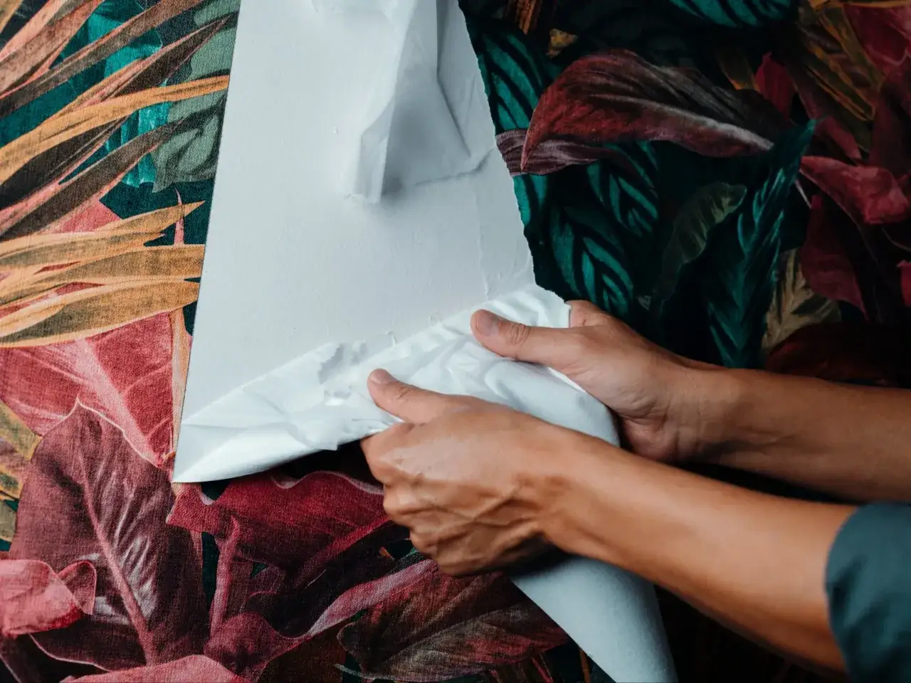 Dłonie odrywają białą warstwę tapety ze ściany z motywem liści. Prosty sposób, jak usunąć tapetę ze ściany.