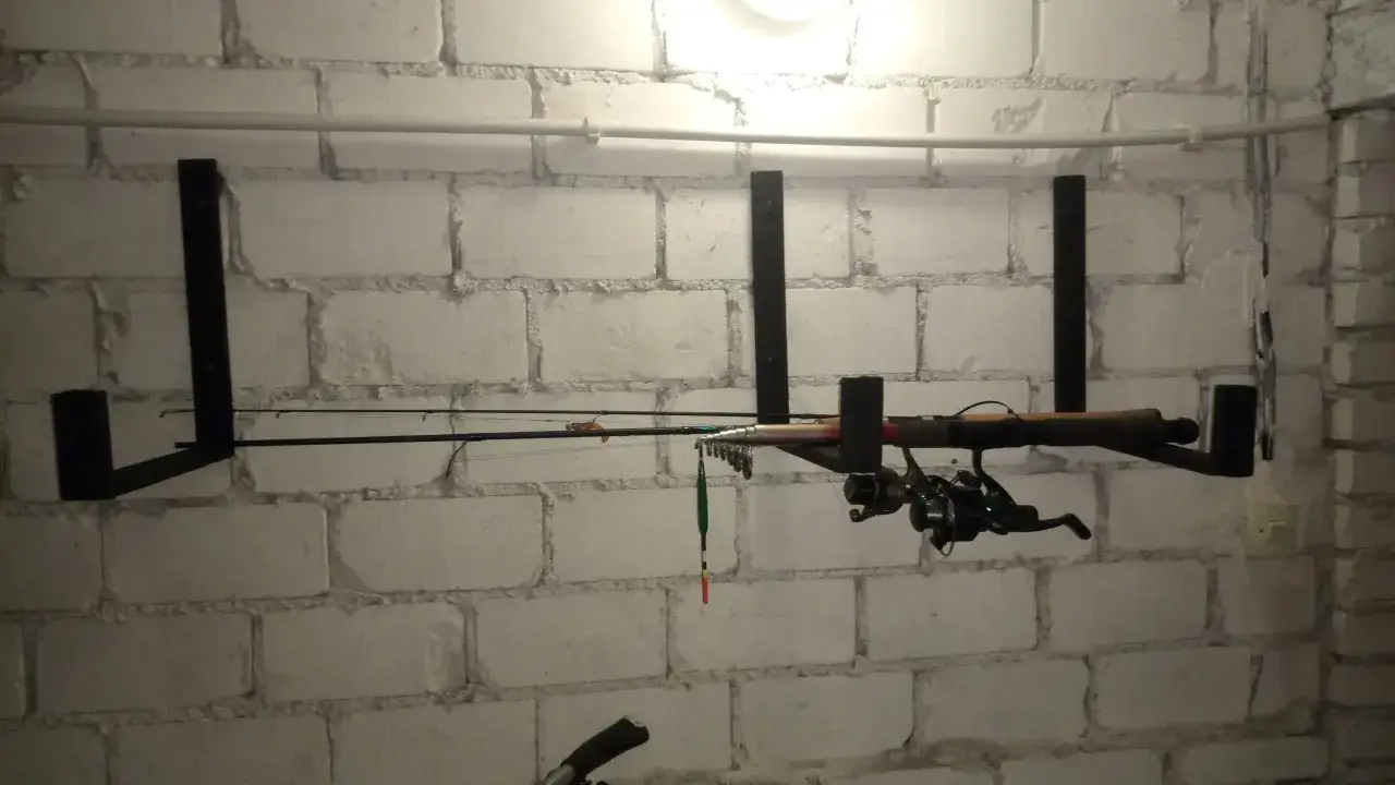 Wieszak na wędki z kołowrotkiem i spławikiem, zamontowany na białej ceglanej ścianie.