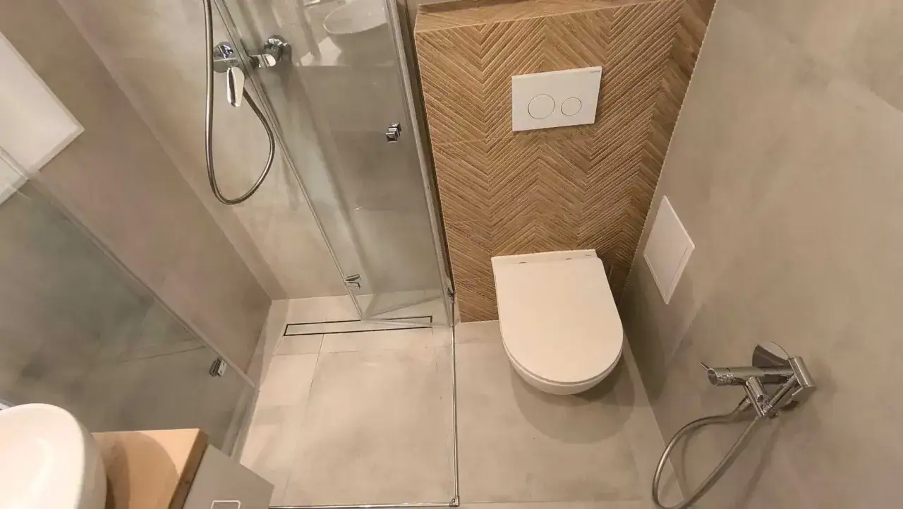 Kompaktowa, bardzo mała łazienka z prysznicem. W rogu biała miska WC, obok niej panel spłuczki z dwoma przyciskami. Ściana za WC ozdobiona drewnianymi płytkami w jodełkę.