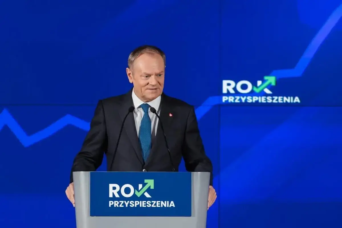 Donald Tusk w garniturze przemawia za mównicą na tle niebieskiej sceny z napisem "Rok Przyspieszenia".